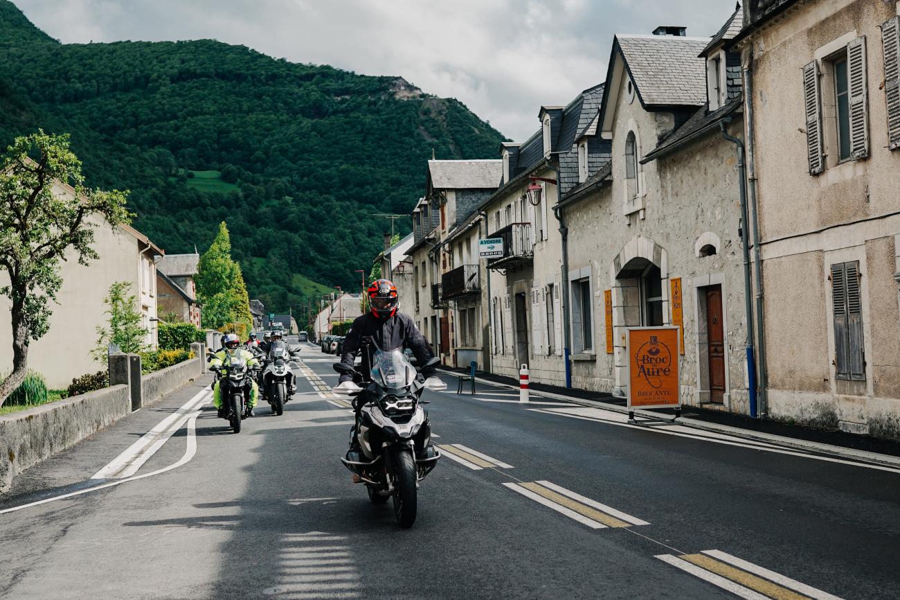 Motorcycle touring group riding through a quaint stone-built French Pyrenees village street.