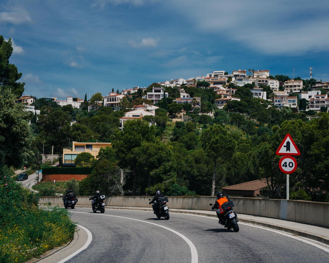 Group of motorcyclists navigating a curving road through a hillside Spanish coastal town.