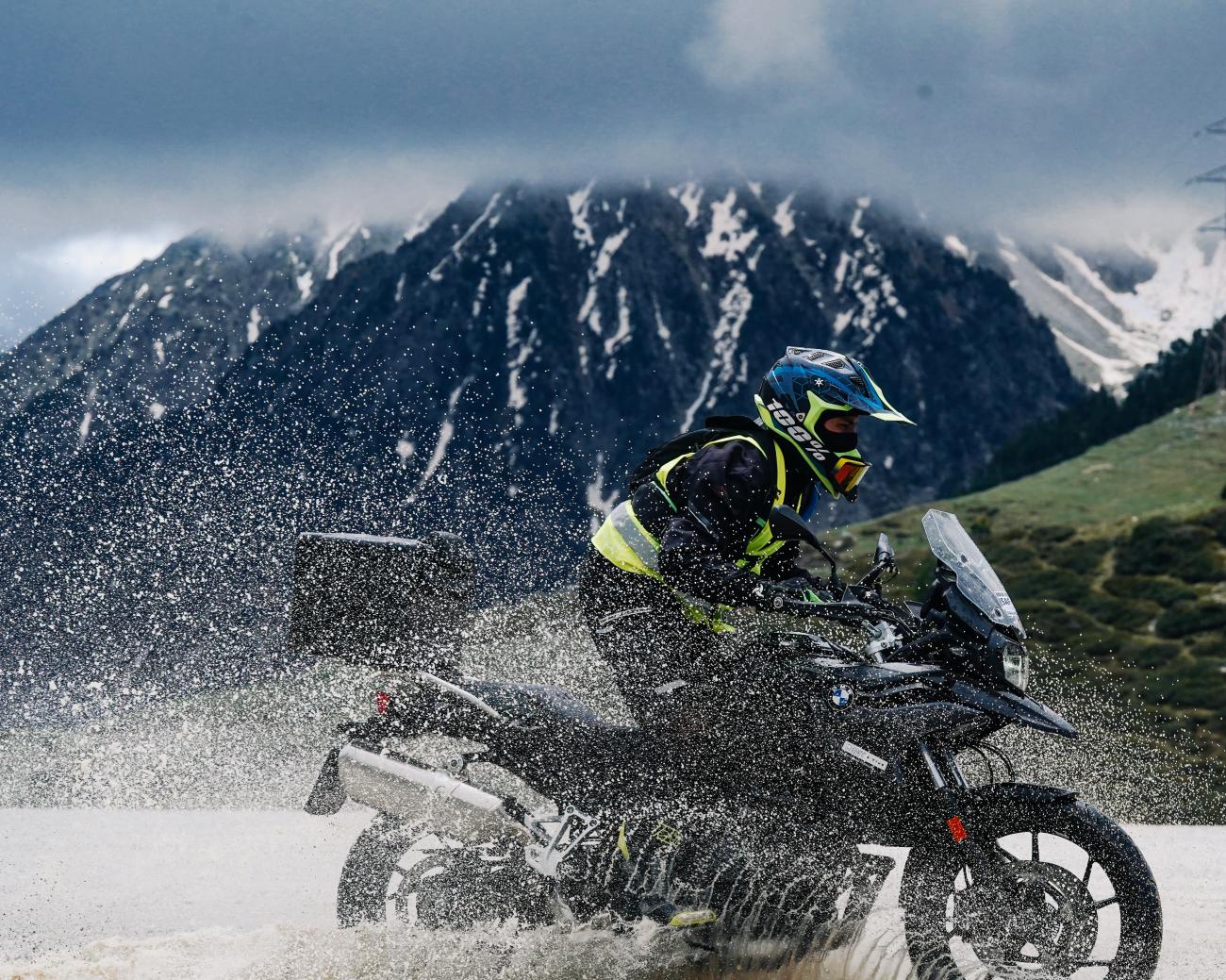 Adventure motorcyclist splashing through water with dramatic snow-streaked Pyrenees mountains in the background.