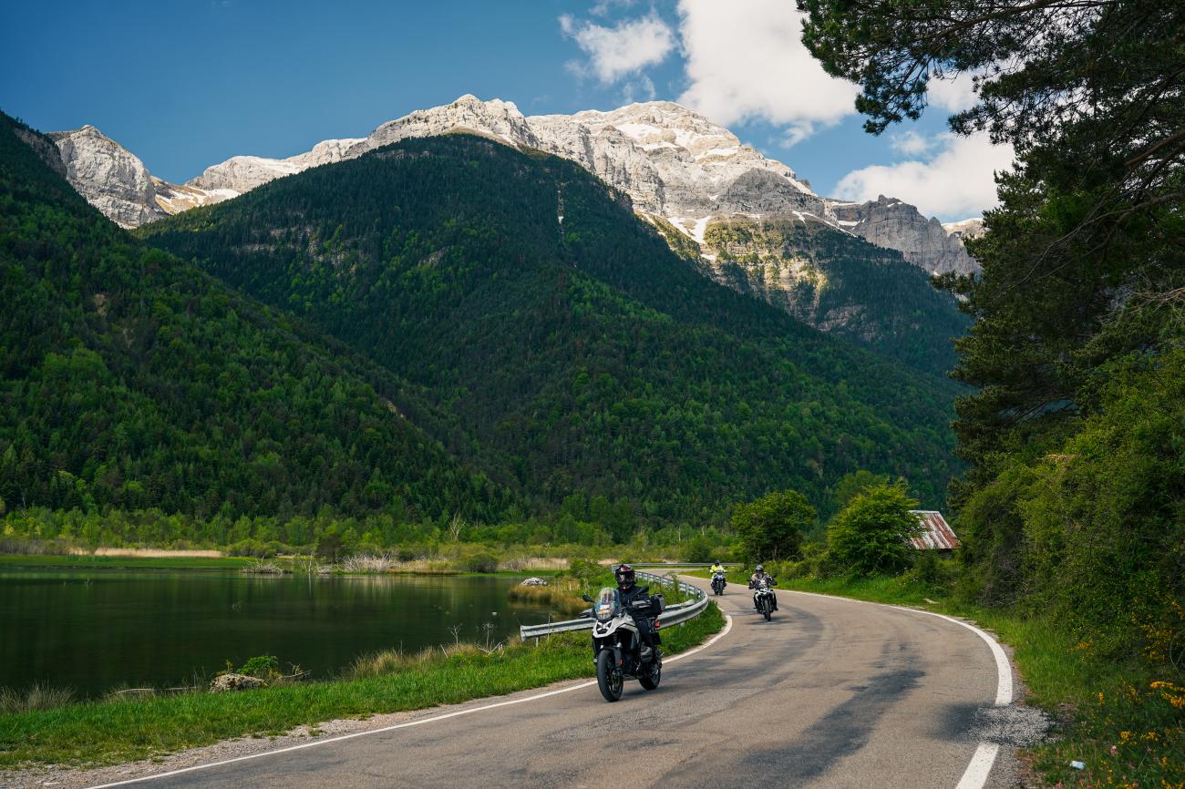 Motorcyclists riding along a lakeside road beneath snow-capped alpine peaks in the Pyrenees.