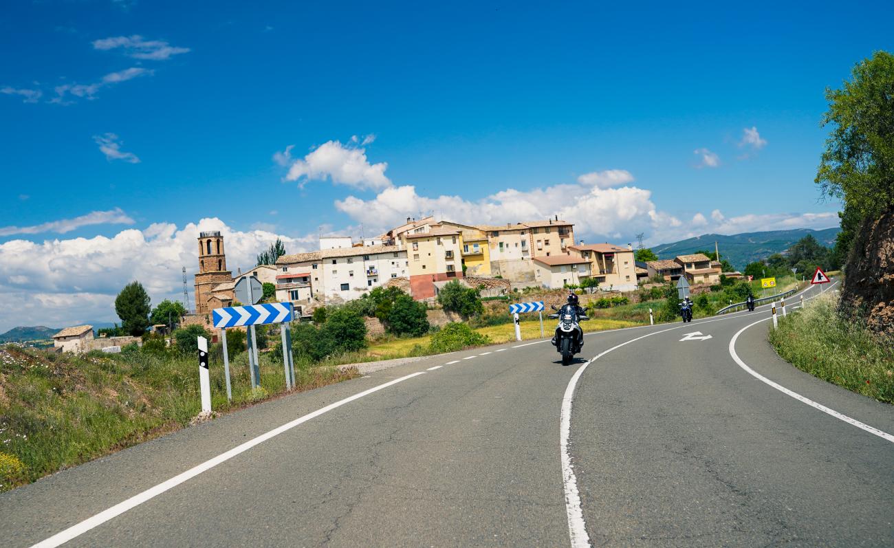 Motorcyclists riding past a historic hilltop village with a church tower on a sunny road in Aragon, Spain.