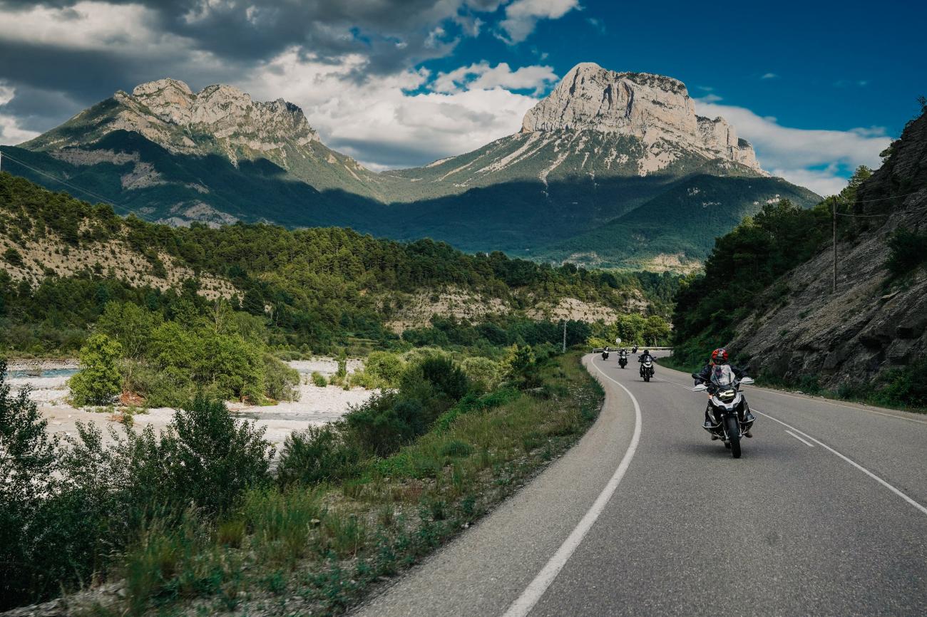 Motorcyclists riding along a scenic Spanish Pyrenees valley road with towering limestone peaks and lush green vegetation.