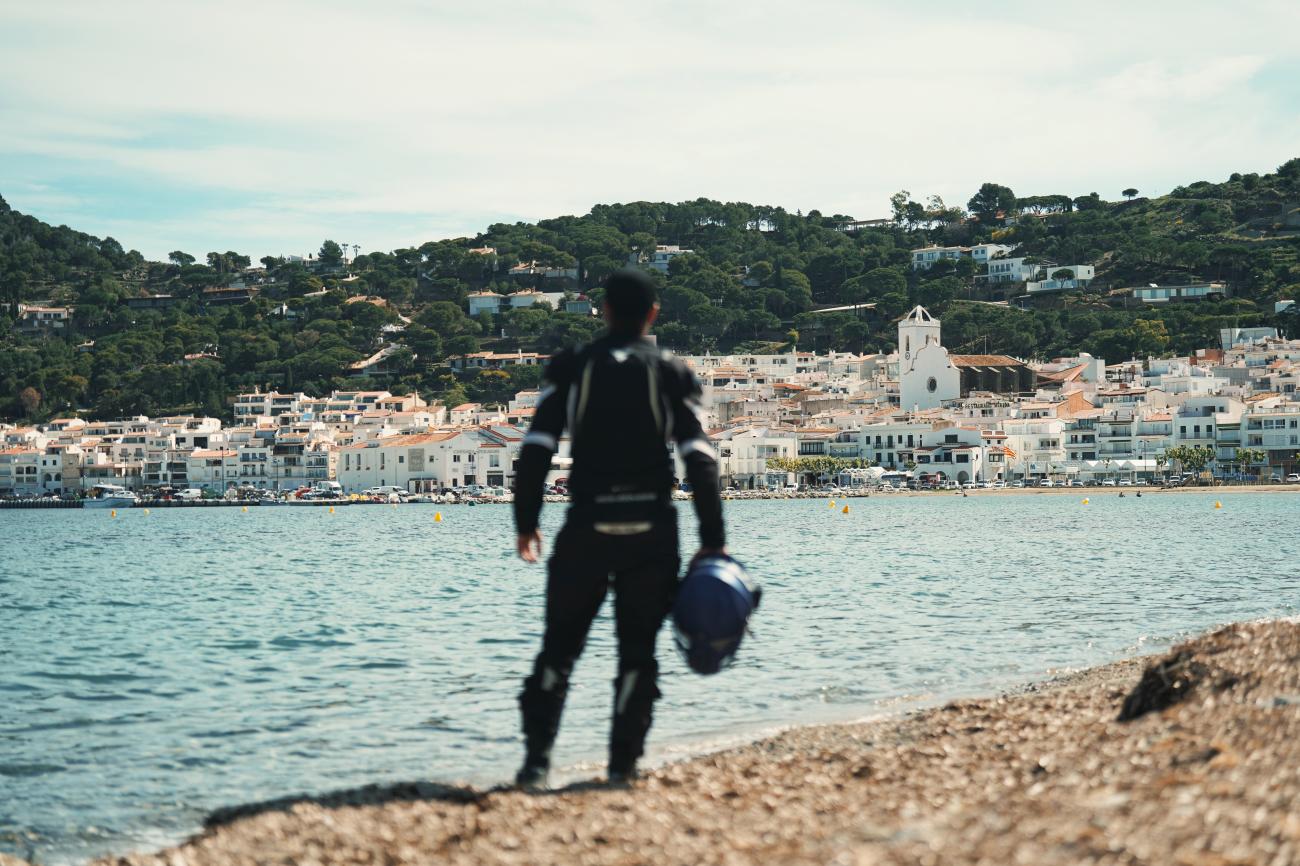 Motorcyclist standing on the beach looking across the bay at a whitewashed Mediterranean coastal village in Spain.