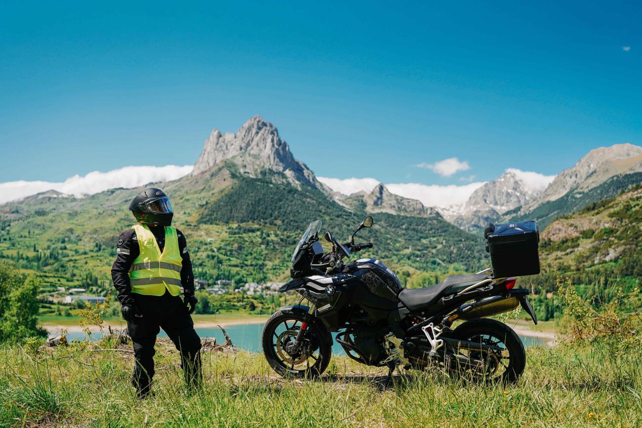 Motorcyclist standing beside a BMW adventure bike overlooking a turquoise Pyrenees lake with jagged mountain peaks behind.