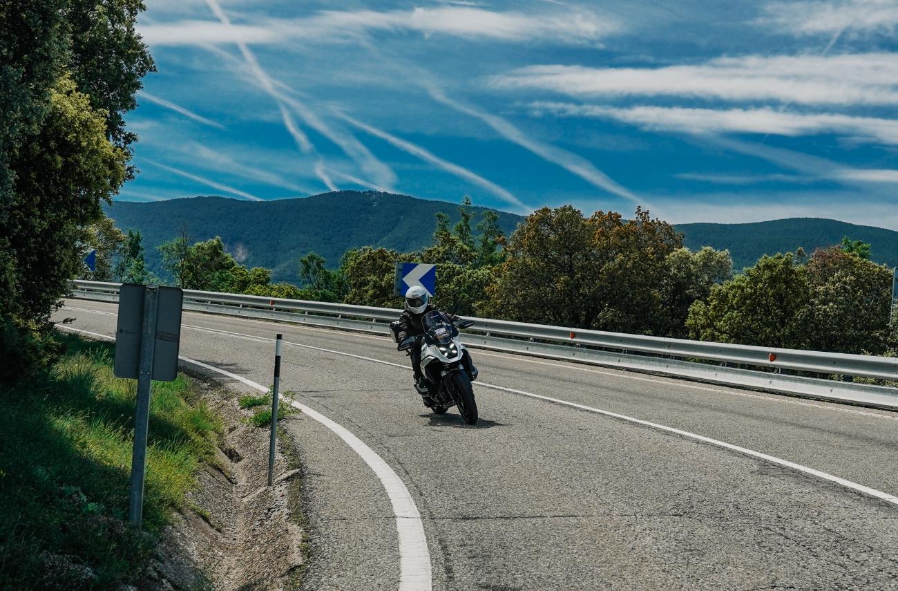 Solo motorcyclist cruising along a sweeping French highway with dramatic mountain range and streaked clouds overhead.
