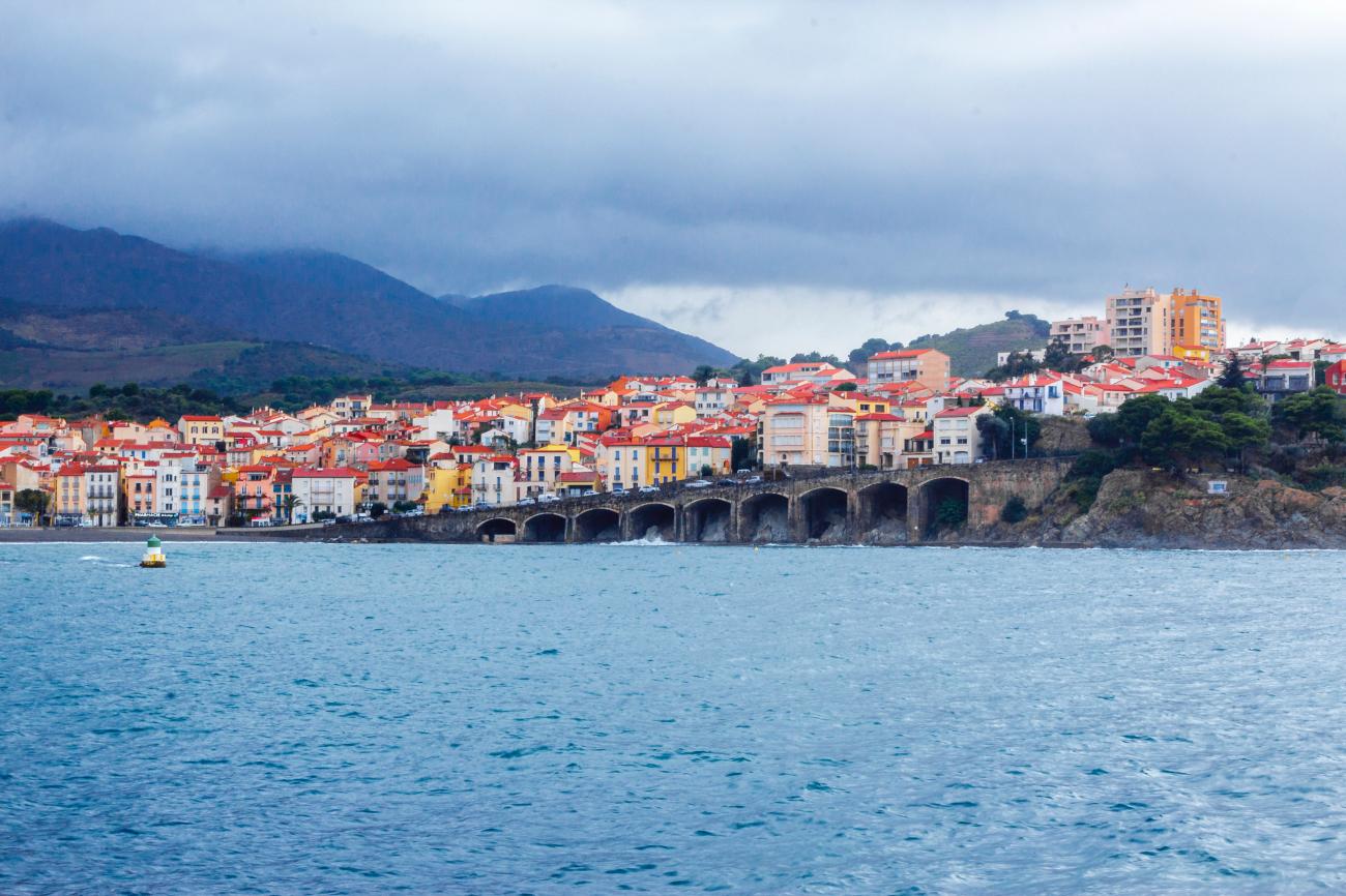Colorful red-roofed houses of a Mediterranean coastal town seen from the sea with misty mountains behind.