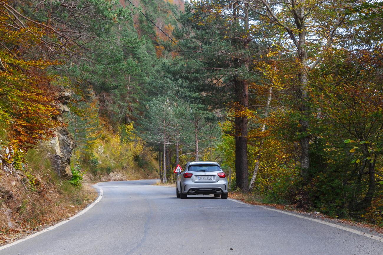 Silver Mercedes driving through a curving road surrounded by colorful autumn forest in the Spanish Pyrenees.