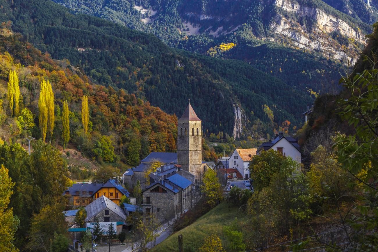 Charming Pyrenees village with a Romanesque church tower nestled among autumn trees with a waterfall on the cliff behind.