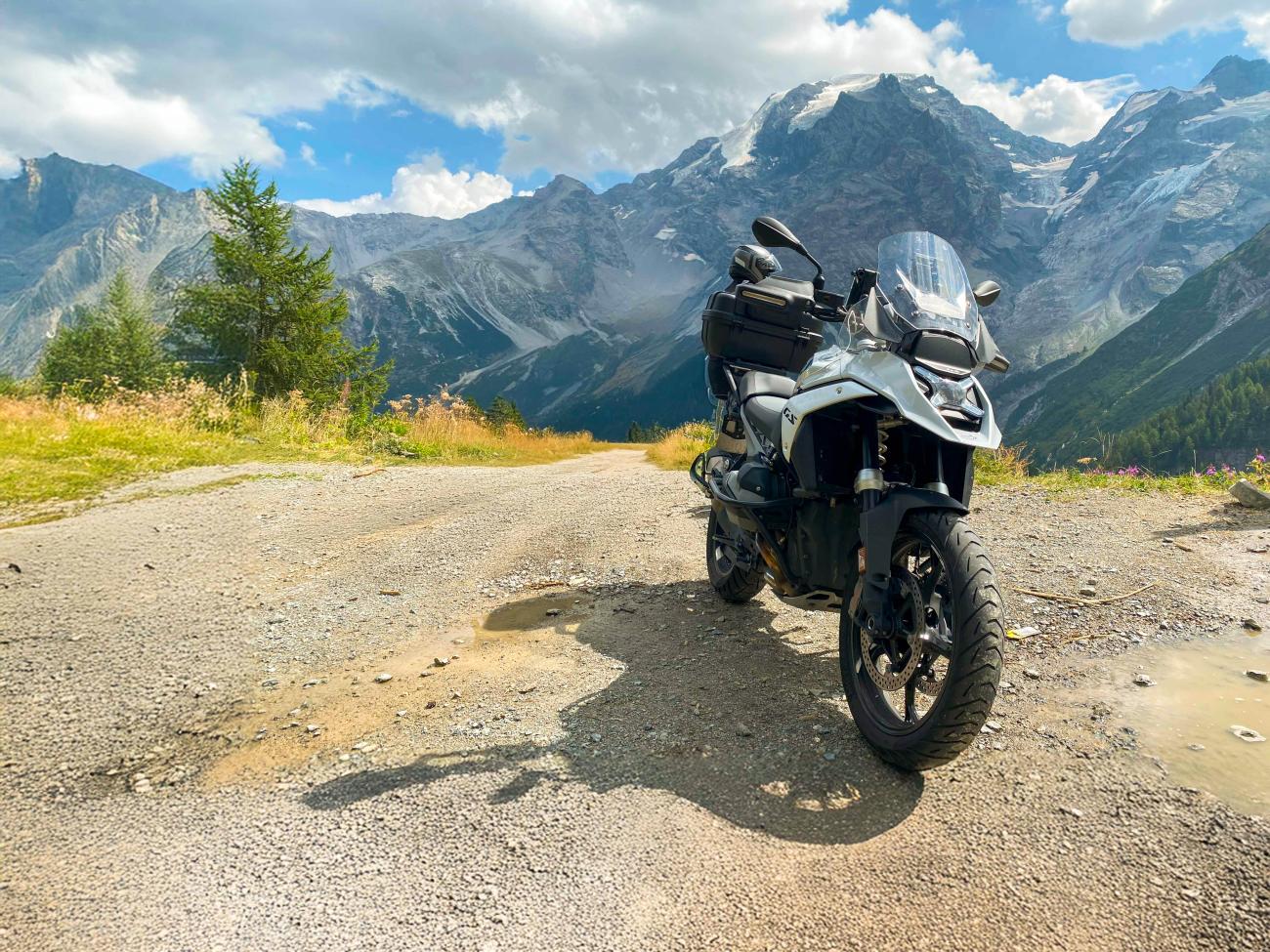 A BMW adventure motorcycle parked on a gravel overlook with a stunning panorama of snow-covered Alpine peaks.