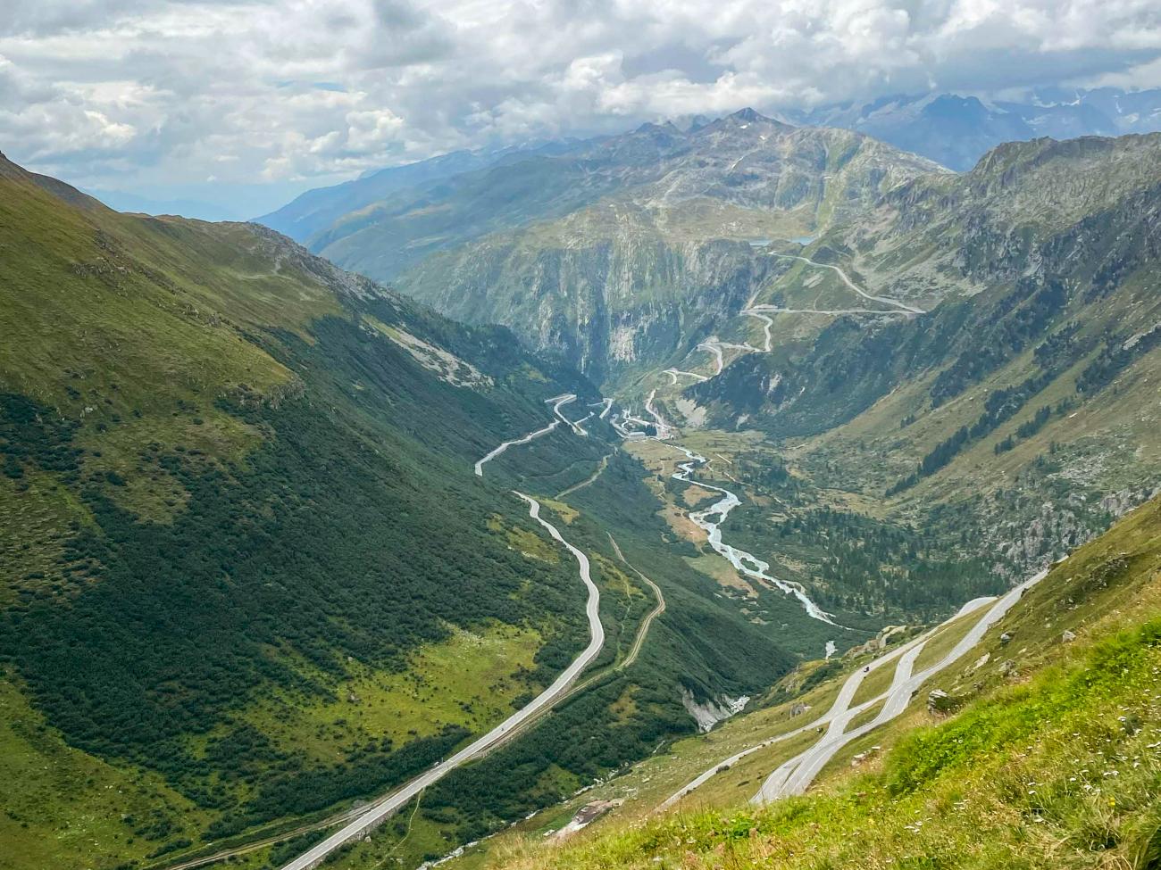 Sweeping view of the Furka Pass valley with a winding river and serpentine roads cutting through green Swiss Alpine slopes.