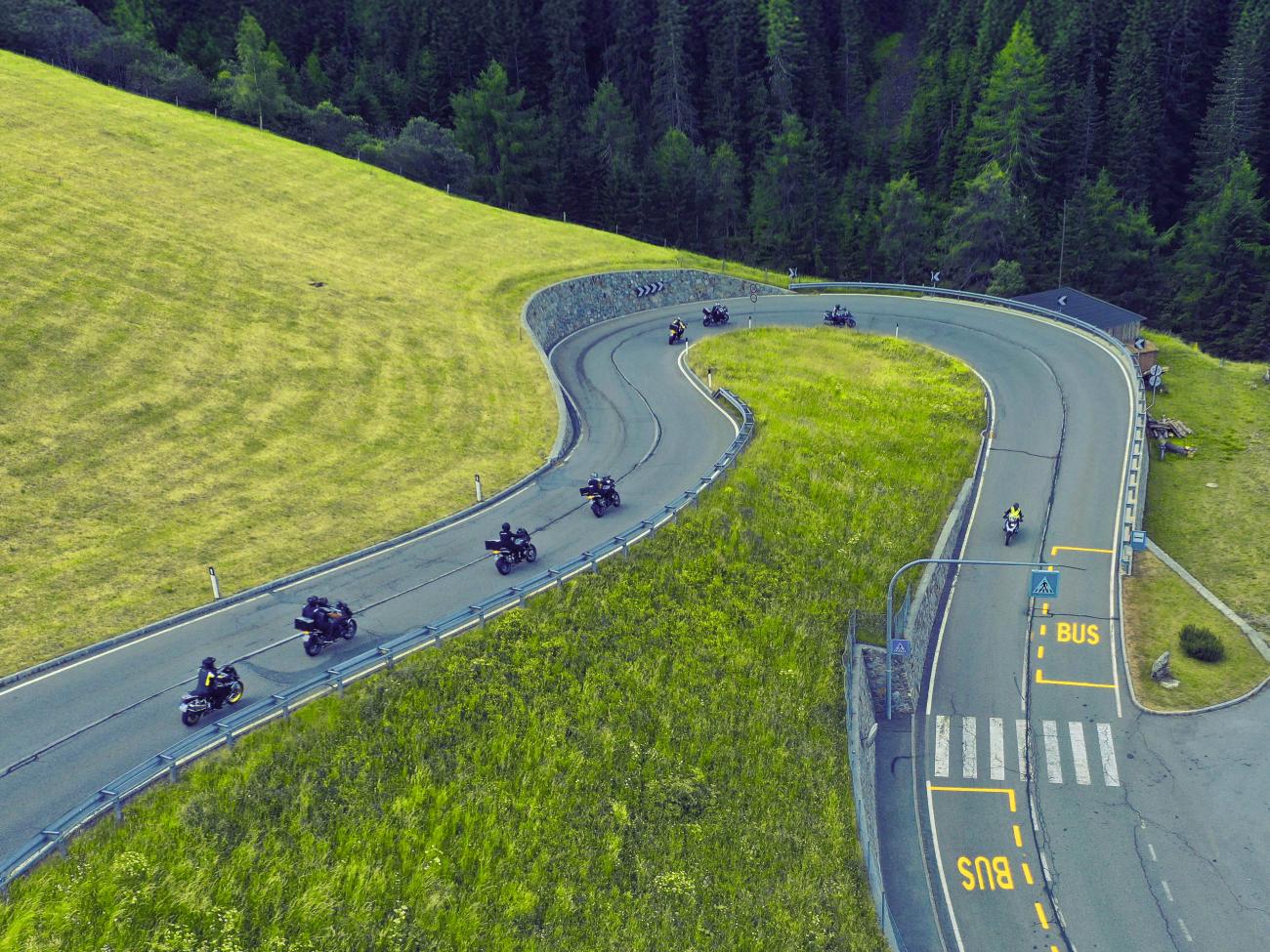 Aerial view of a group of motorcyclists riding through a sweeping curve on a lush green alpine road.